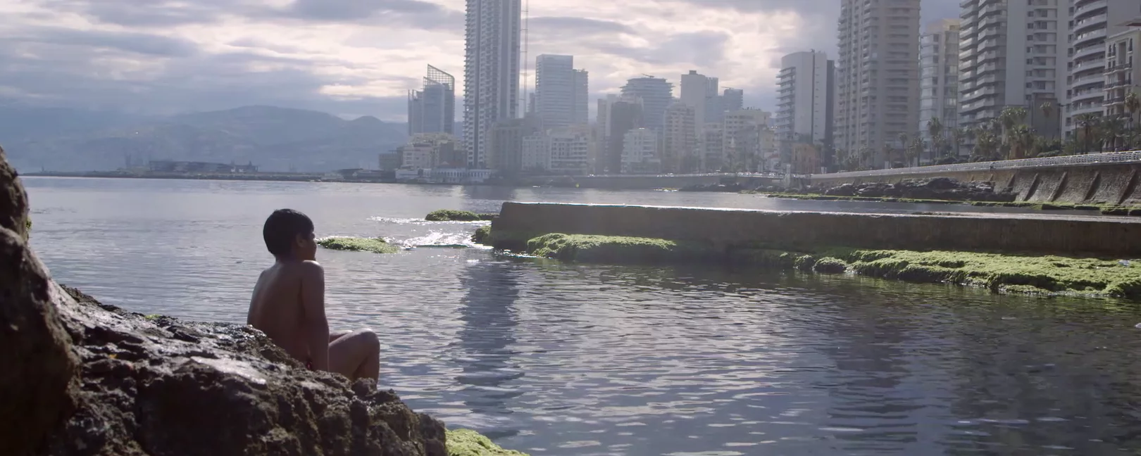 Boy sitting on the riverside with the city appearing on the other bank