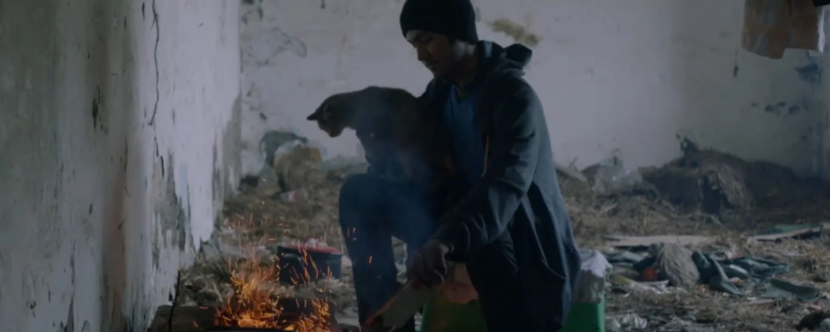 A young man holding a cat and getting worn of fire in what appears to be ruins of an abandoned house