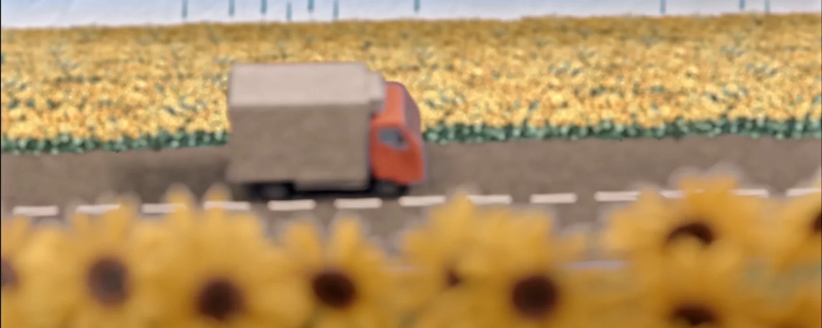 Still from animation movie. The Still shows a truck on a road stretching between sunflower fields