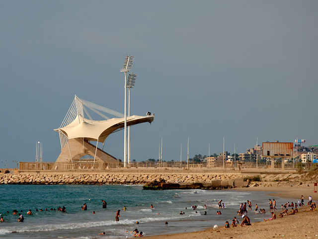 The stand of Saida International Stadium with a well occupied (and slightly rubbish strewn) beach in the foreground